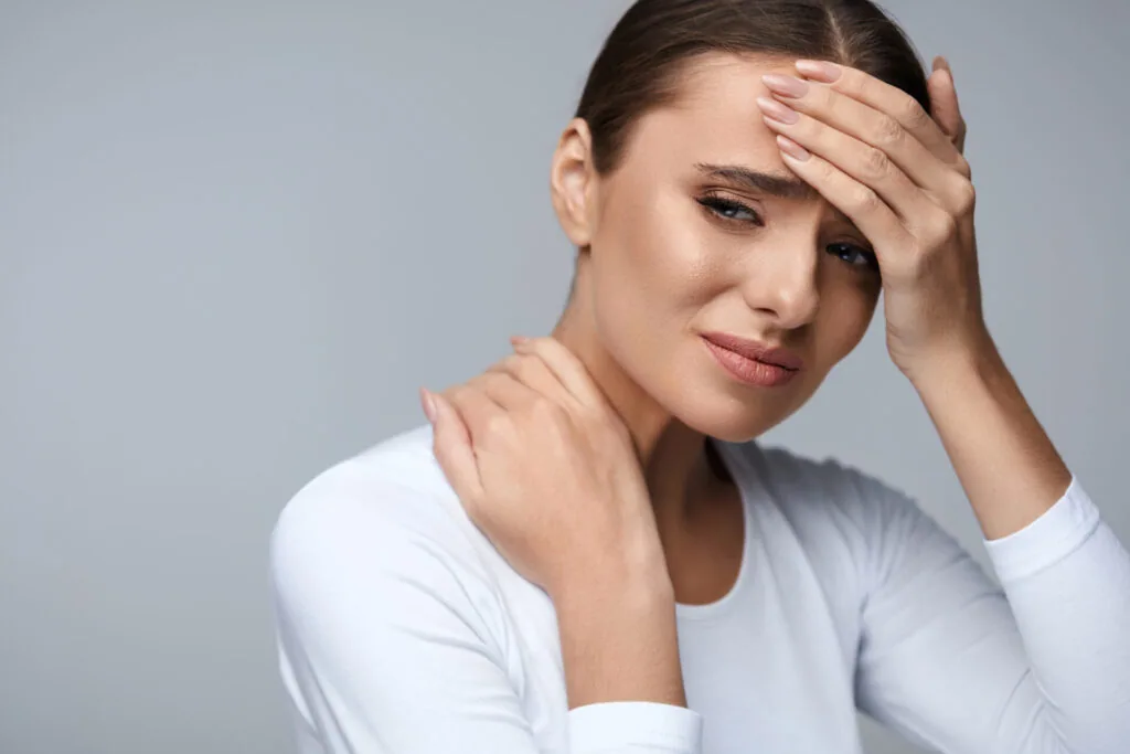 Woman holding her neck and forehead in discomfort against a gray background