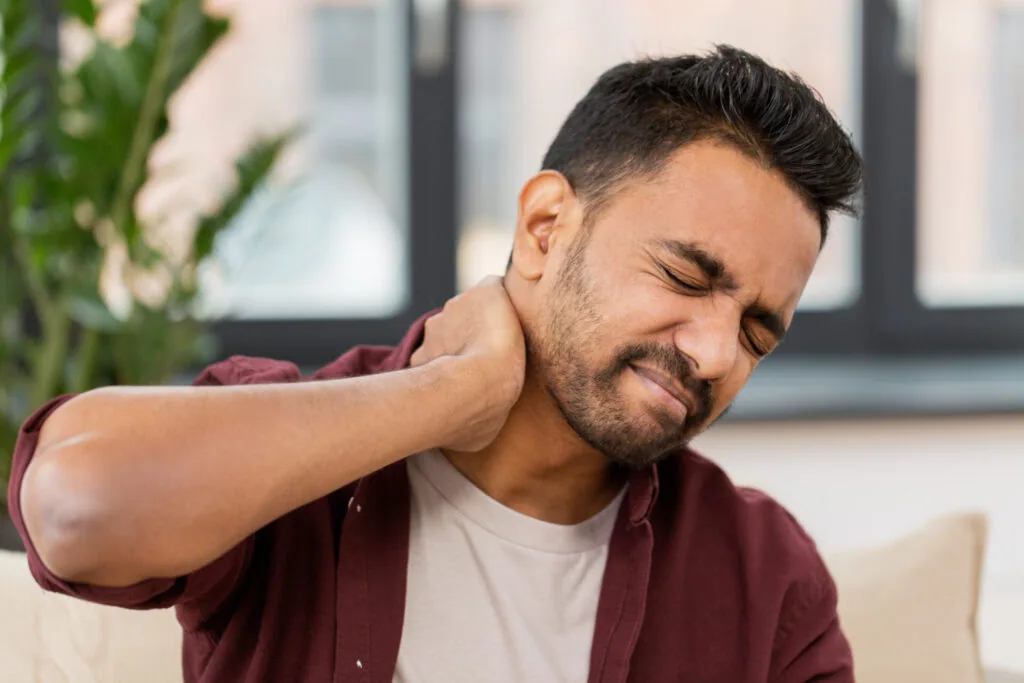 Man in maroon and white shirt touching his neck with eyes closed, grimacing in discomfort indoors