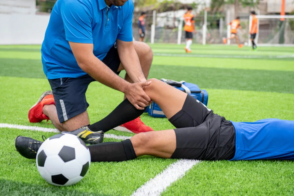 Athletic trainer examining injured soccer player's knee on field while other players practice in background