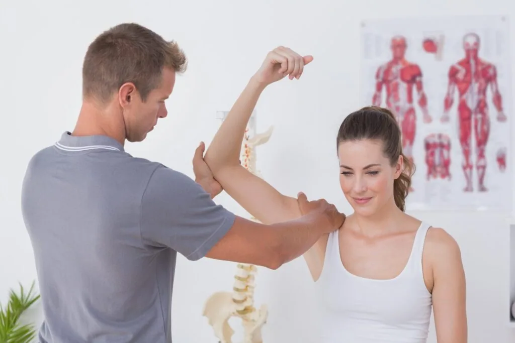 Physical therapist examining patient's arm flexibility in clinical setting with anatomical charts on wall